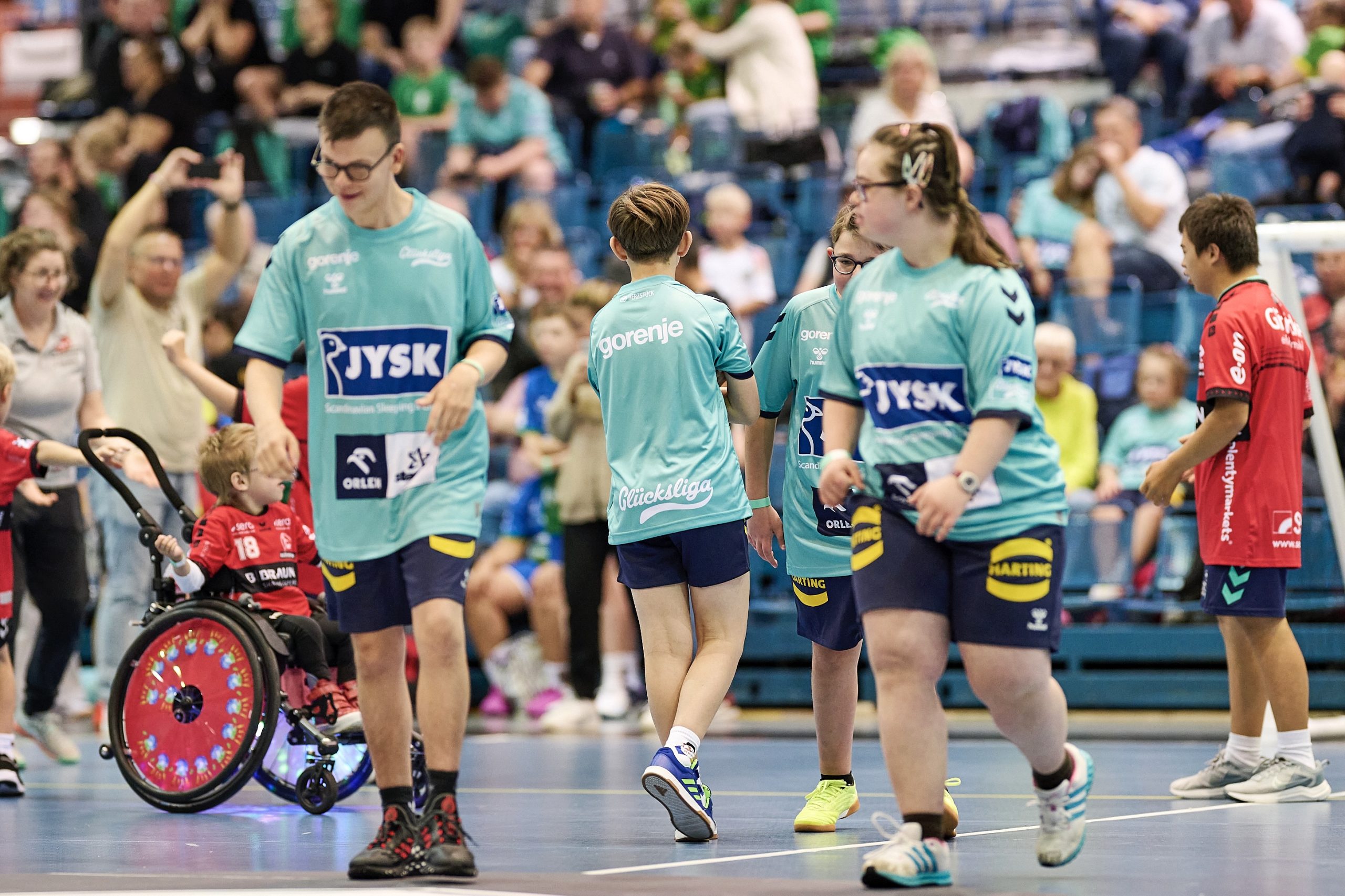 Kinder in Glücksliga-Trikots gehen über ein Spielfeld in einer Sporthalle